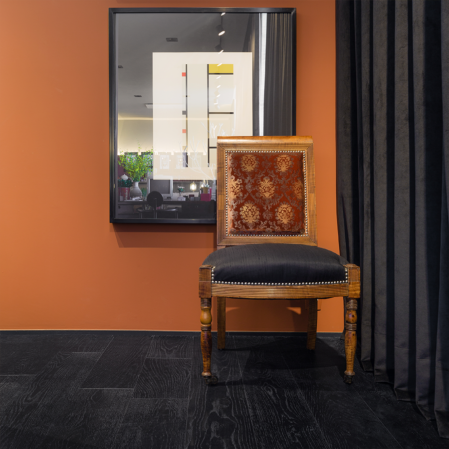 Fotografia de parede, com quadro que reflete o ambiente no vidro e cadeira decorativa posicionada no canto, cortina preta de cetim na lateral e piso de madeira preto.