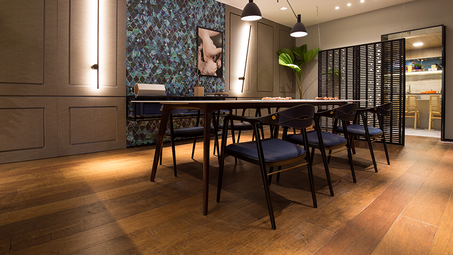 Imagem de uma sala de jantar com piso de madeira Multistrato, uma mesa e seis cadeiras, parede com luminárias compridas e um quadro. Ao fundo, uma planta e uma divisória que leva a uma área com mesa e cadeiras.