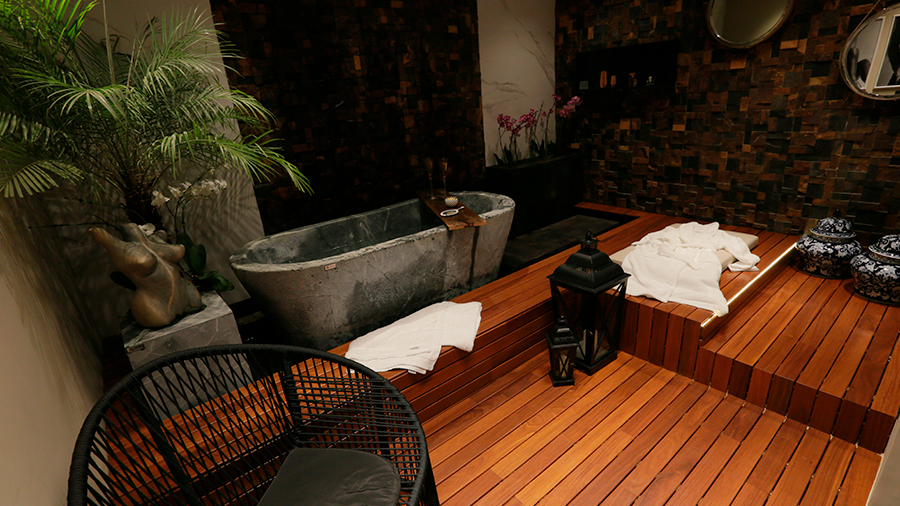 Fotografia de um banheiro com deck de madeira, banheira de concreto, toalhas, plantas e objetos de decoração
