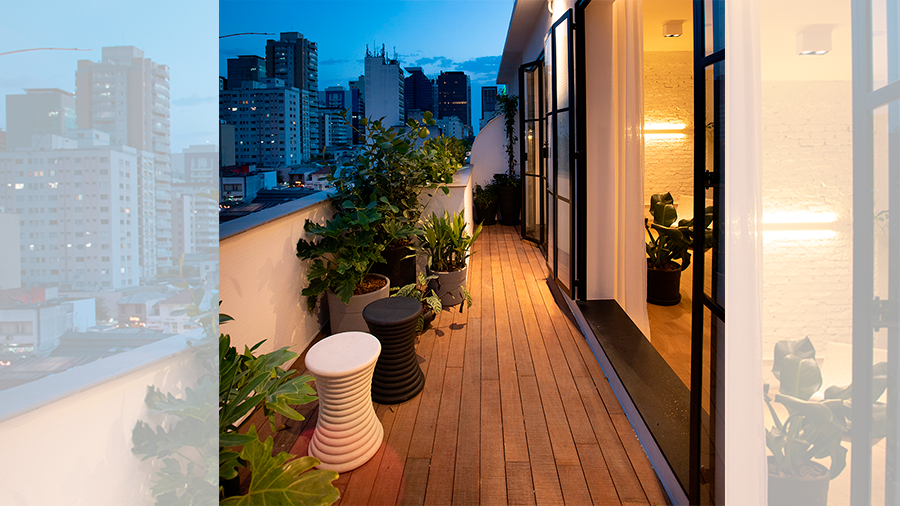 Fotografia de uma varanda com deck de madeira, vasos de plantas, bancos e uma vista para prédios
