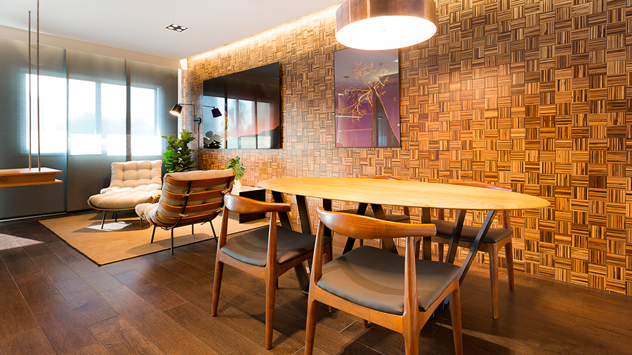  Imagem de um ambiente com mesa oval de madeira, quatro cadeiras e um lustre. Ao fundo, há um tapete com duas poltronas e quadros. A parede tem revestimento de madeira Multiestruturado.