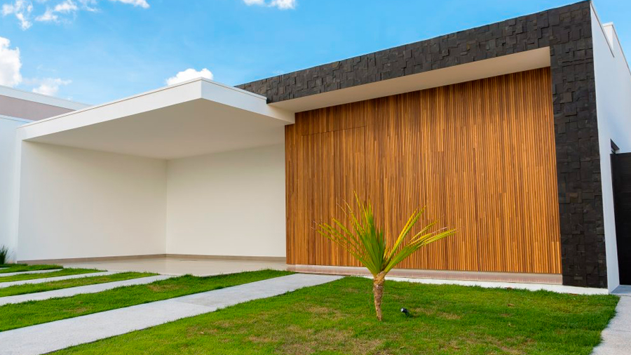 Imagem da fachada de uma casa branca e preta com metade de uma parede de madeira Brise Cumaru; na frente, grama e uma árvore plantada.