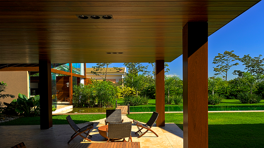 Área externa com cadeiras e mesa de centro de madeira, coberta por um teto de madeira. Ao redor, há grama, plantas e diversas árvores.