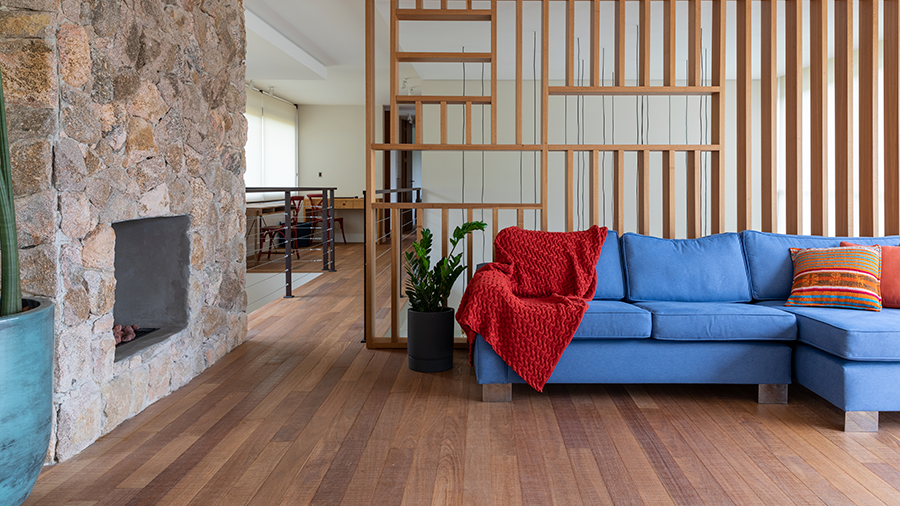 Sala com assoalho de madeira cumaru de demolição, sofá com manta e almofadas encostado em um ripado de madeira, vaso ao lado e, na parede lateral, uma lareira.