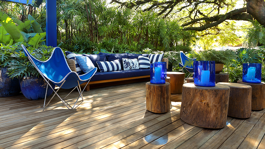 Imagem de uma área externa com deck de madeira, cilindros de madeira com vasos azuis em cima. Do outro lado, um sofá com assento e encosto em azul royal, almofadas listradas, e uma cadeira retrátil ao lado com plantas e árvores ao redor.