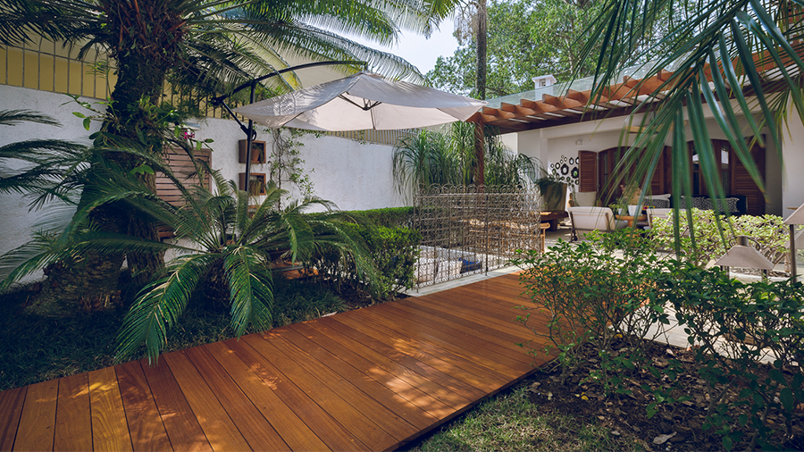 Imagem de um quintal com coqueiros e outras plantas. No centro, há um caminho feito com deck de madeira Cumaru que termina na entrada de uma casa branca com janelas marrons.