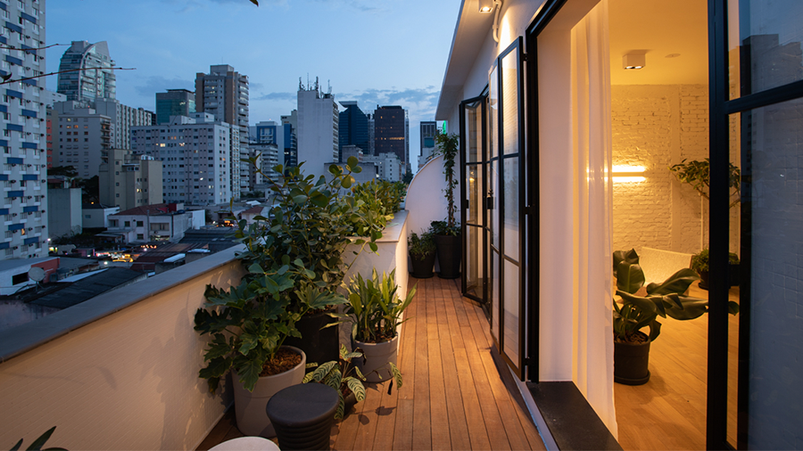 Imagem da varanda de um apartamento com deck de madeira Cumaru. Na área, há vasos com plantas, portas de vidro com estrutura preta, e na lateral, o ambiente interno do apartamento com a luz acesa.