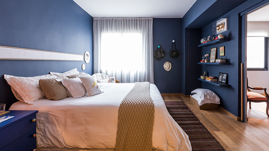 Imagem de um quarto com paredes azuis, uma cama arrumada em tons de branco e marrom, prateleiras com objetos, piso de madeira Cumaru e um tapete.