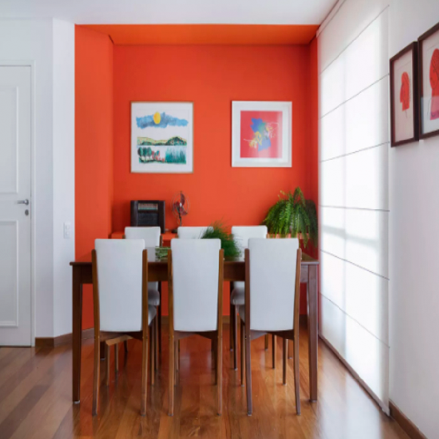 Foto da sala de jantar com parede única laranja, contrastando as demais paredes brancas, quadros na parede e presença de plantas, alguns objetos decorativos e uma cortina na porta da sacada, o piso é de madeira.