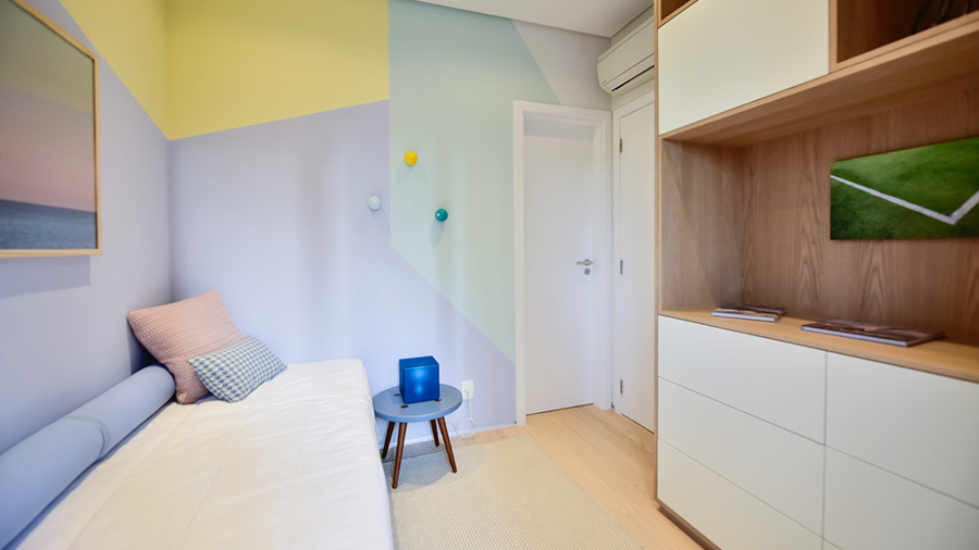 Imagem de um quarto com cama e almofadas claras, um banco ao lado e um armário de madeira à frente. As paredes apresentam cores claras e padrões geométricos.