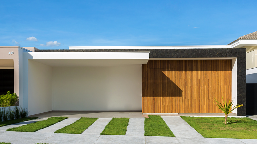 Imagem externa de uma casa com formato quadrado, sendo metade branca e metade revestida com madeira Brise. Na frente, há piso e grama.