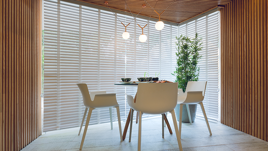 Imagem de uma sala minimalista com uma janela que se abre para uma mesa cercada por três cadeiras brancas, onde há utensílios sobre ela. Acima, há um lustre, e ao lado, um vaso com uma planta. As paredes são revestidas de madeira Brise.