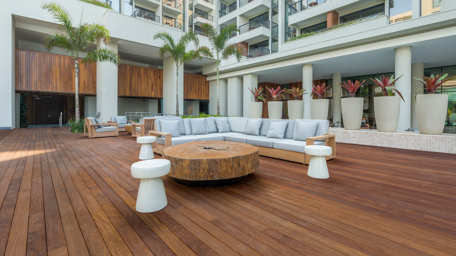 Área de lazer de edifício, com piso de madeira, decorada com palmeiras e vasos de planta. Ao centro, mesa, bancos e sofás