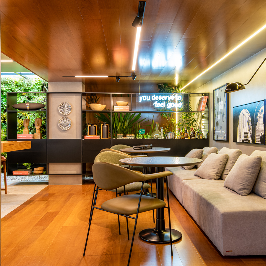  Imagem de um ambiente com piso e teto de madeira MasterTeto Champagne, contendo um sofá, mesa e cadeiras. Na parede, quadros, e ao fundo, prateleiras exibindo diversos objetos decorativos e plantas.