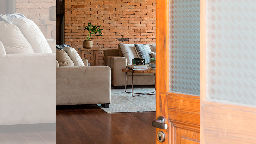 Fotografia de um espaço interno com piso de madeira, sofá, tapete, parede de tijolinho, mesa de centro e objetos de decoração.