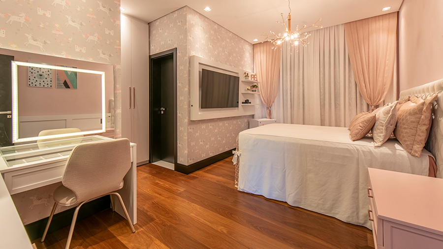  Fotografia de um quarto com piso de madeira, cama de solteiro, papel de parede, penteadeira, televisão e cortinas em cores claras.