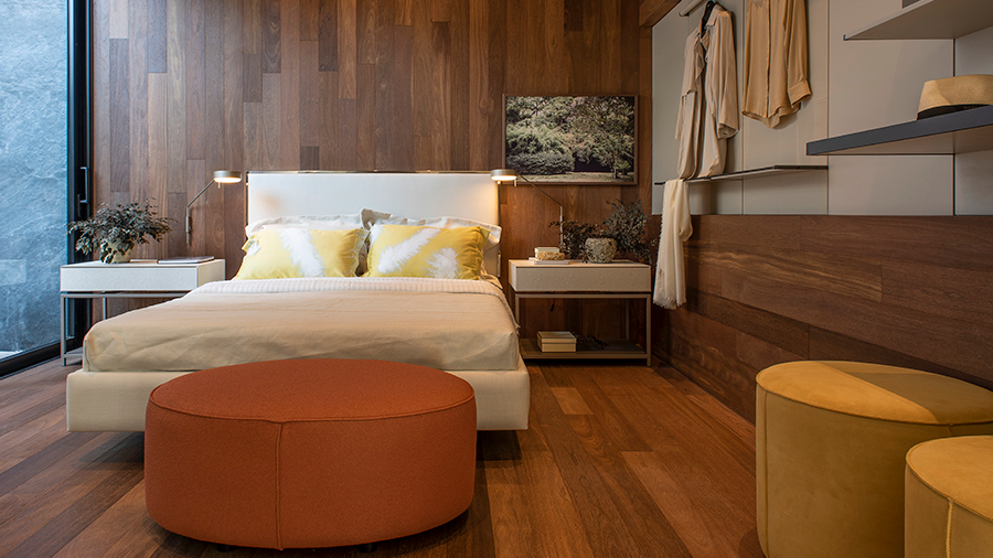 Fotografia de um quarto com piso de madeira, cama de casal, mesas de cabeceira, pufes e itens de decoração.