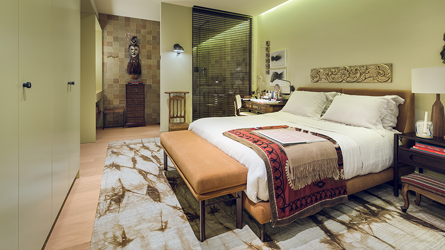 Fotografia de um quarto com piso de madeira, cama de casal, banco, tapete estampado, mesas de cabeceira e itens de decoração.