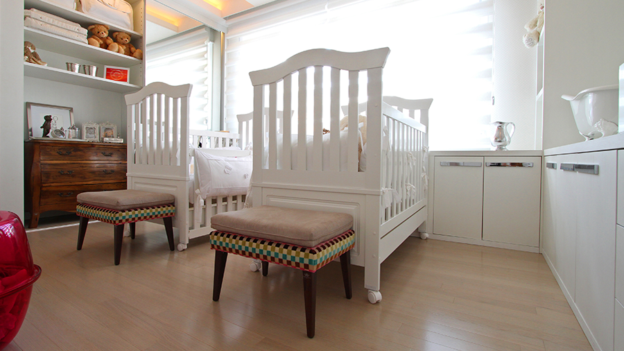 Fotografia de um quarto de bebê com piso de madeira, berço branco, banco, armários e itens de decoração.