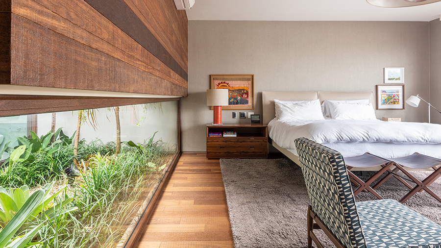 Fotografia de um quarto com piso de madeira, cama de casal, tapete bege, mesa de cabeceira, luminárias, itens de decoração e grande janela com vista para jardim.