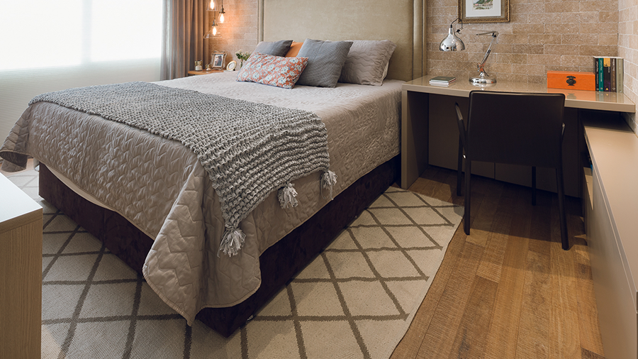 Fotografia de um quarto com piso de madeira, cama de casal, tapete estampado, escrivaninha com cadeira, almofadas e itens de decoração.
