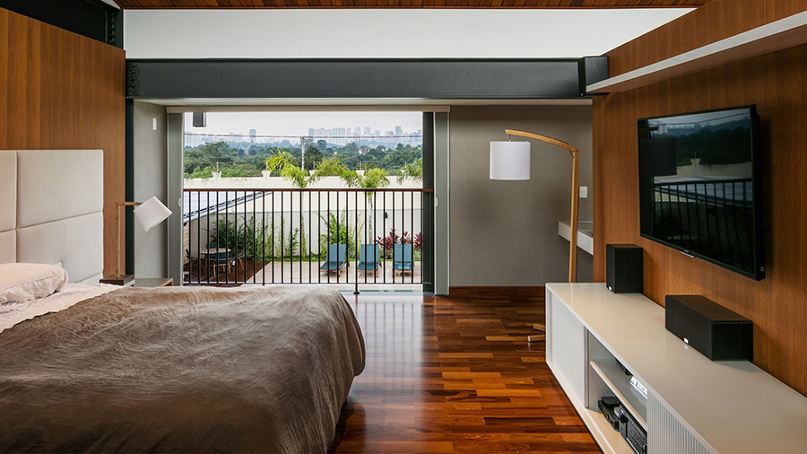 Fotografia de um quarto com piso de madeira, cama de casal, televisão, rack branco e vista para uma varanda externa.