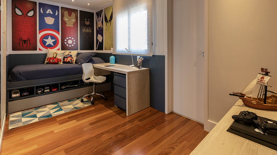 Fotografia de um quarto com piso de madeira, cama de solteiro. escrivaninha, cadeira e posteres de super heróis.