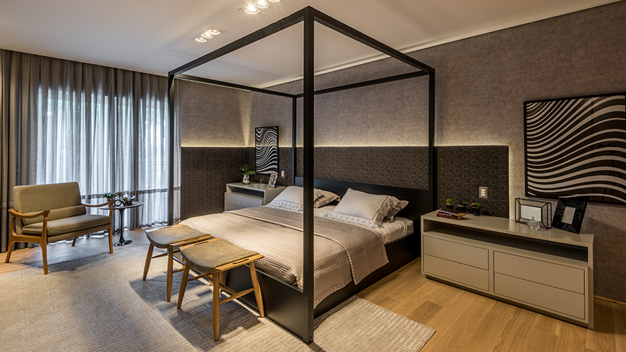 Fotografia de um quarto com piso de madeira, cama de casal, cadeira, poltronas, tapete cinza e mesas de cabeceira.