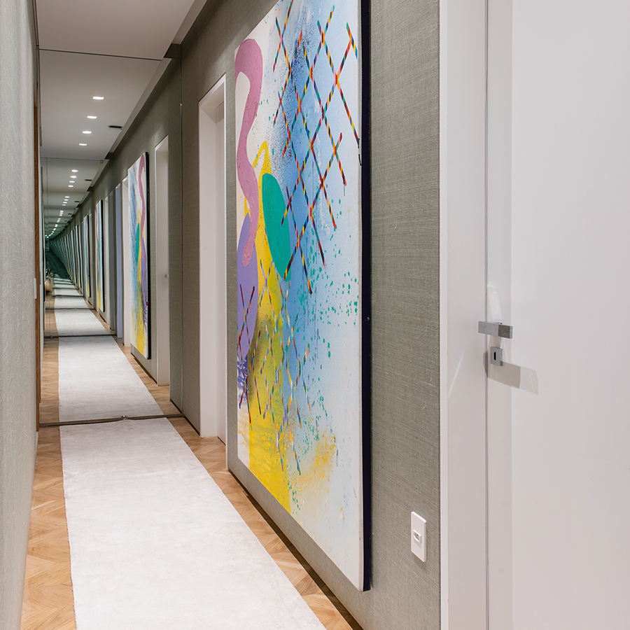 Fotografia do corredor do apartamento de Silvia Braz, piso de madeira, tapete, quadro na parede que ocupa a parede quase inteira entre as portas dos ambientes e um espelho no final do corredor que dá a sensação de profundidade.