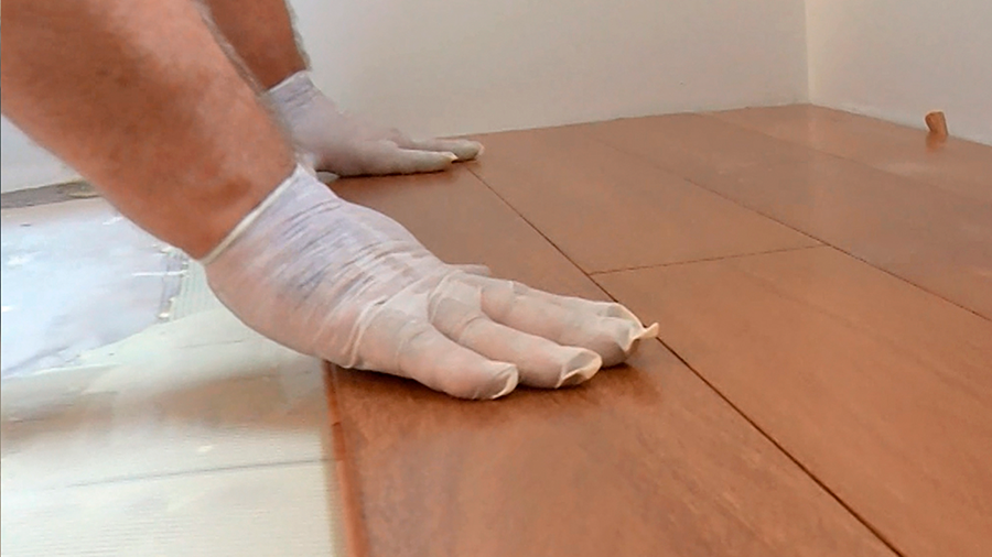 mãos com luvas brancas pressionam placas de piso de madeira contra o chão de azulejo para instalação. 