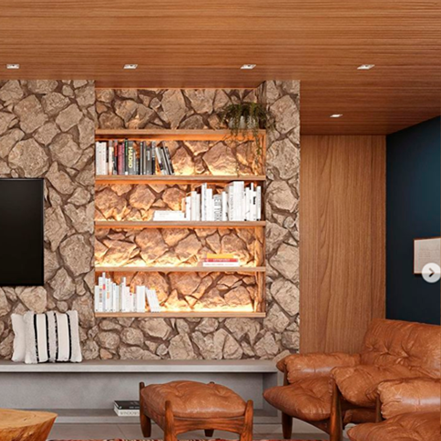 Sala com parede de pedras, estante com livros iluminada, móveis rústicos de madeira e tapete artesanal colorido.