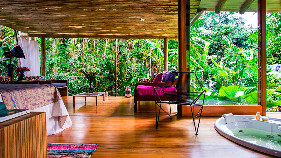  Fotografia de interior de casa integrada a natureza, com piso de madeira, decoração colorida e paredes em vidro.