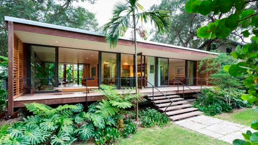  Fotografia de ambiente externo de casa tropical, com paredes em vidro, revestimentos em madeira e jardim ao redor.