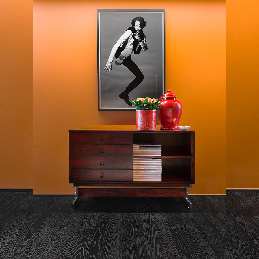 Fotografia de um espaço interno com piso de madeira escura, móvel de madeira, parede na cor laranja, quadro com imagem em preto e branco e vasos de plantas.
