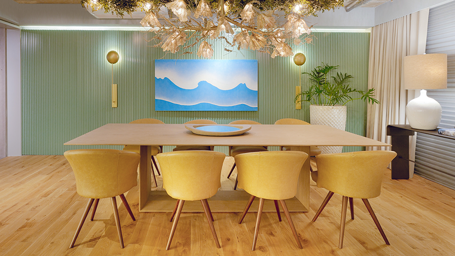 Fotografia de uma sala de jantar com piso de madeira, lustre em formato de plantas, parede verde, mesa de madeira e poltronas amarelas.