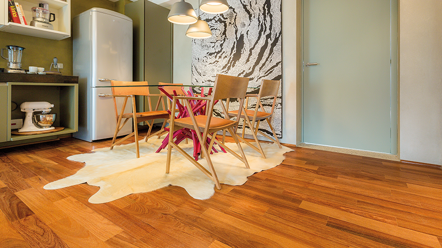 Fotografia de uma cozinha com piso de taco de madeira na cor escura, tapete, mesa, itens de decoração e eletrodomésticos.