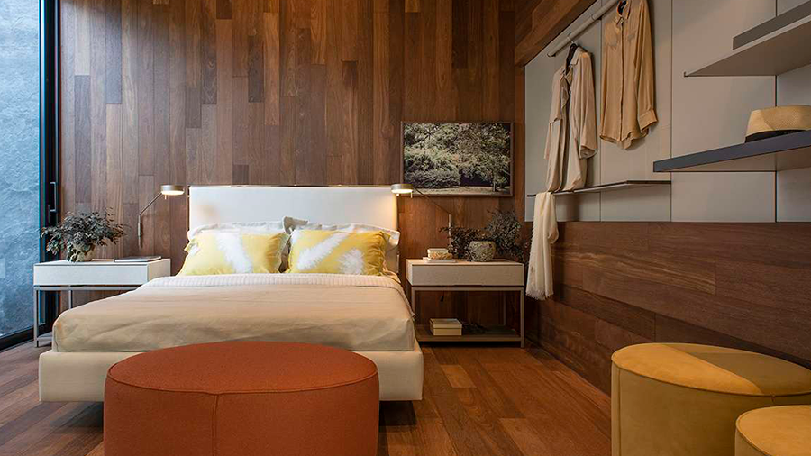 fotografia de um quarto com acabamentos em madeira, cama e assentos centralizados e porta de vidro lateral. 