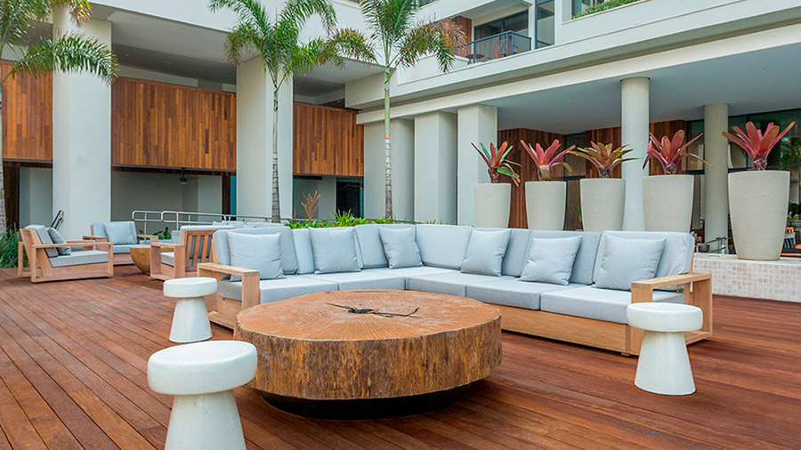 deck de madeira com mesa central e sofás, ao redor estão vasos e árvores tropicais. 