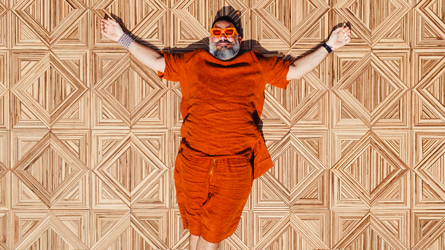 Imagem do designer Pedro Franco com roupas e óculos laranja deitado no piso Ancestralidade de madeira.
