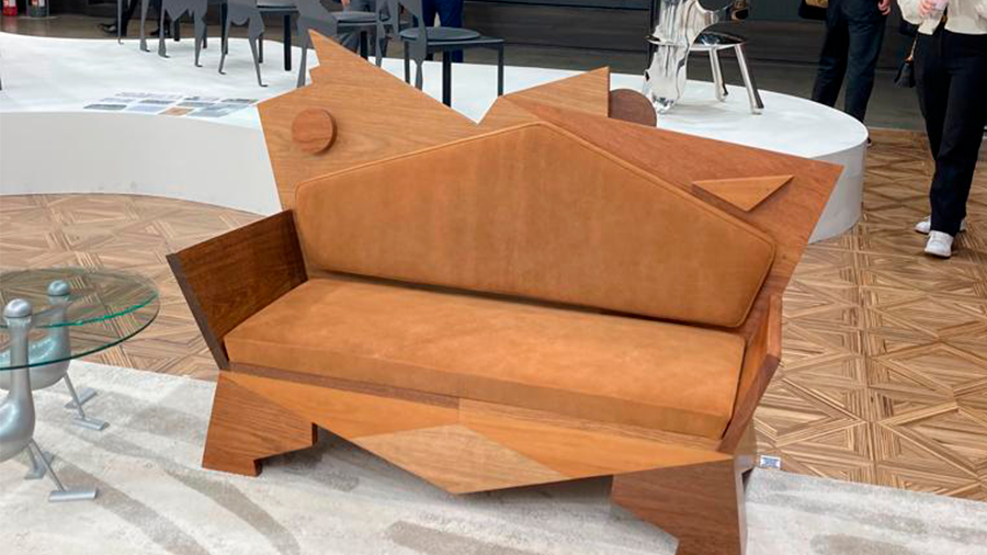 Imagem do sofá K2 assimétrico de madeira no Salão do Móvel em Milão, rodeado por um piso de madeira, uma mesa de centro de vidro e pessoas. Ao fundo, há móveis em exposição sobre um palco.