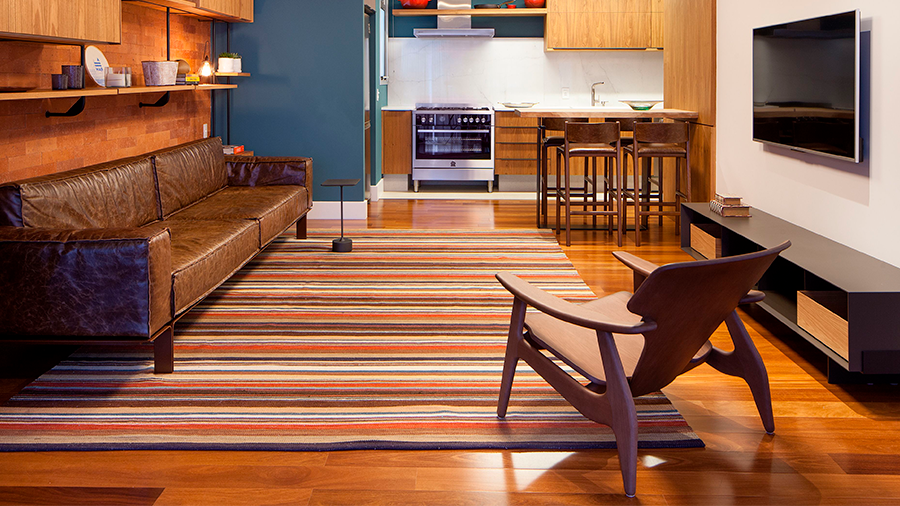 sala integrada com cozinha, sofá de couro a esquerda, prateleiras de madeira acima, tapete nas cores vermelho e marrom, cadeira e painel de televisão à frente. Ao fundo, cozinha com armários azuis. 