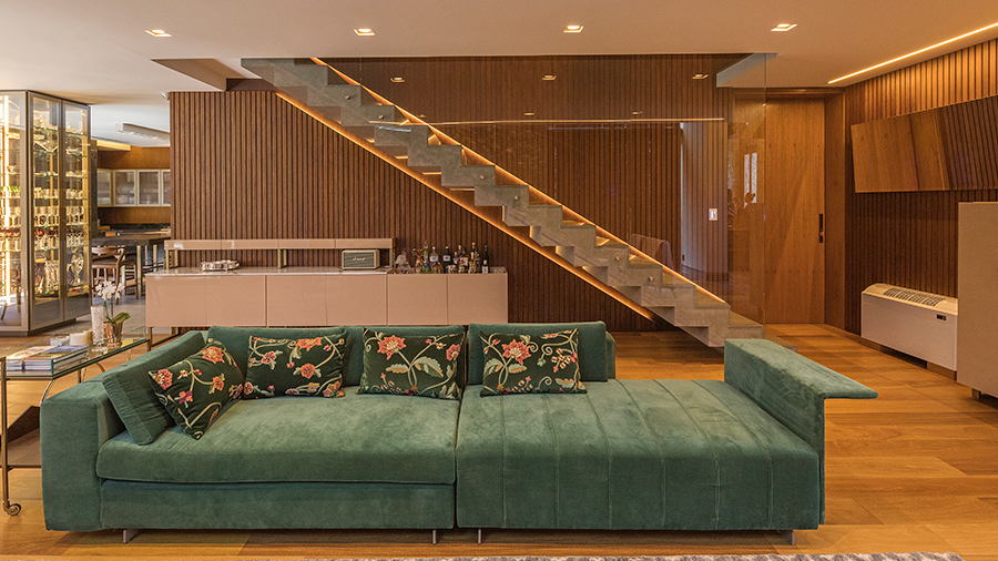 Salva de estar com piso e revestimento de madeira nas paredes, sofá grande verde no centro e escada ao fundo
