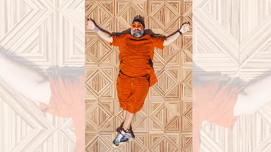 Fotografia de um homem vestindo peças de roupa laranja deitado de braços abertos sobre um piso de madeira.