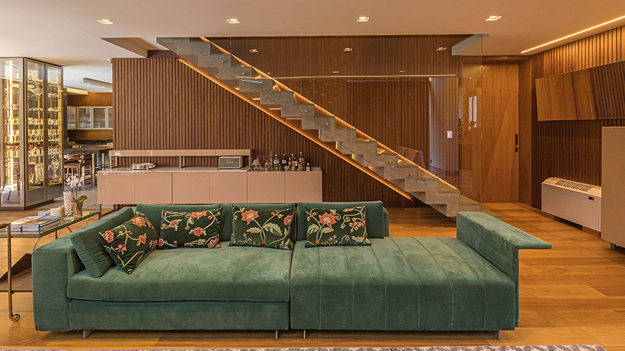 Fotografia de um uma sala de estar com piso de madeira, sofá verde, amofada, móveis, escada, tapete e objetos de decoração