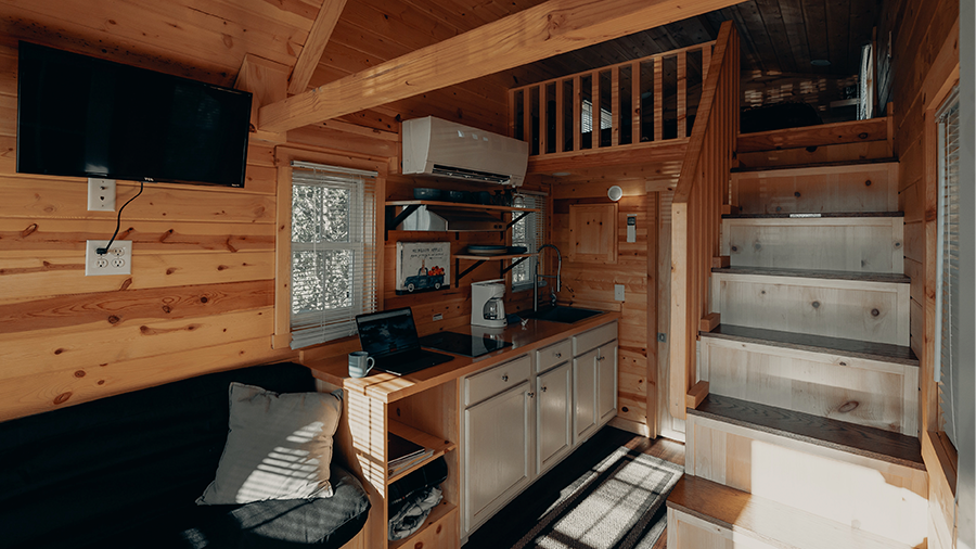 Fotografia do interior de uma casa com paredes, escadas e toda a estrutura de madeira, móveis, utensílios de cozinha, eletrodomésticos, sofá preto e uma almofada cinza.
