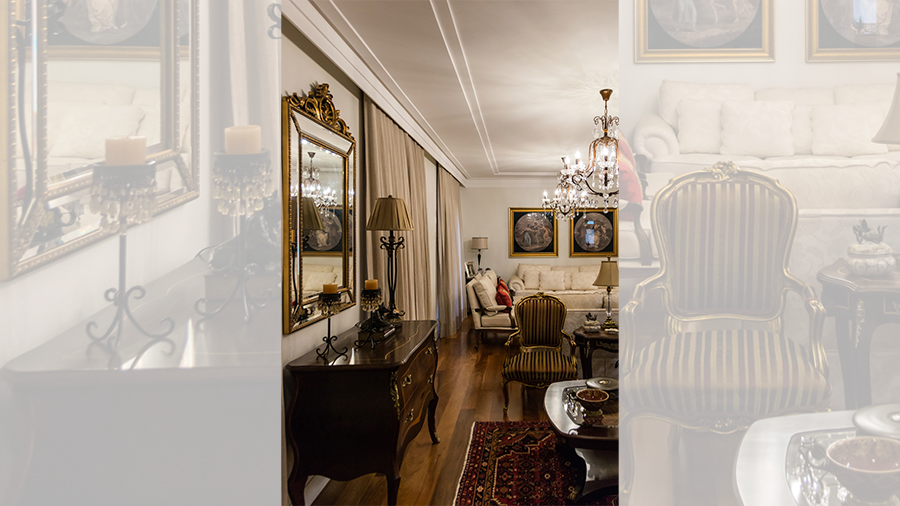 Fotografia de um espaço interno com piso de madeira,móveis antifos de madeira, lustre, espelho na parede e itens de decoração.