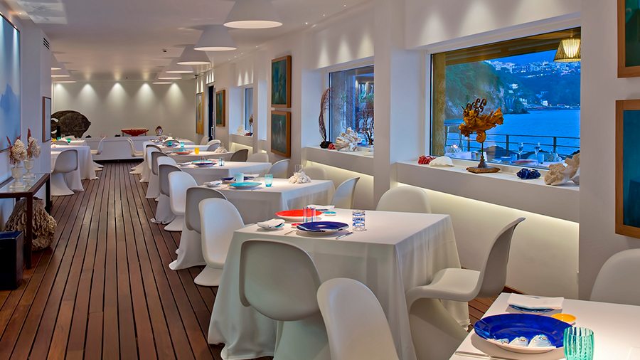 Fotografia de um espaço de restaurante de hotel com piso de deck de madeira, mesas de jantar com toalhas e cadeiras brancas, louça colorida e objetos de decoração.