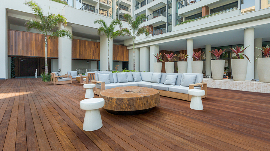 otografia de um espaço externo de hotel com piso de deck de madeira, sofá com almofadas, mesa de centro de madeira rústica e bancos brancos.