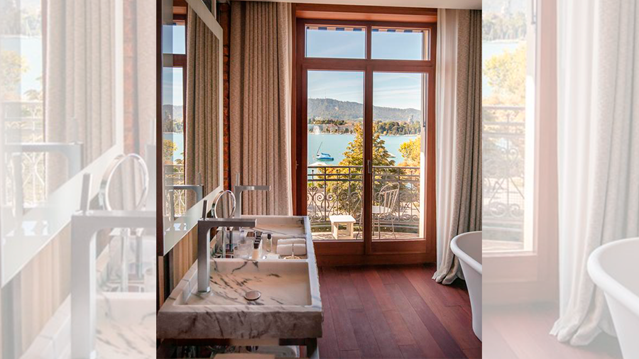 Fotografia de um banheiro de hotel com piso de madeira, pia de mármore e vista para varanda.