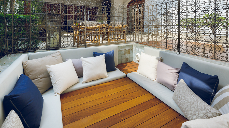 Área externa com divisória de ferro em arabescos, antiga piscina, coberta com deck de madeira e sofá com almofadas revestem todo o entorno